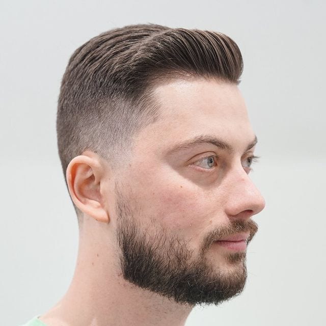 Mens haircut 4 featuring a tiny quiff hairstyle on a guy with a fade and a beard against a grey background