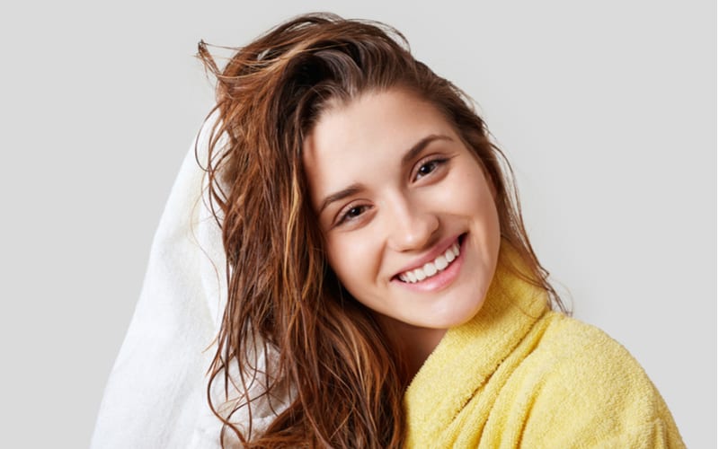 Lovely female with wet hair, takes shower, dries head with towel, being pleased after taking bath, dressed in yellow bathrobe, poses against white background, has cheerful expression
