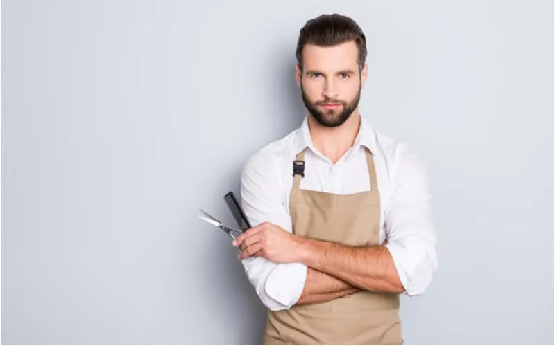 Portrait with copy space, empty place of virile harsh barber having his arms crossed, holding equipments in hand, looking at camera, isolated on grey background for a piece on what is a hair salon