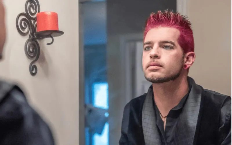 Image of a guy with spiked and dyed hair looks in the mirror and stands next to a candle on the wall