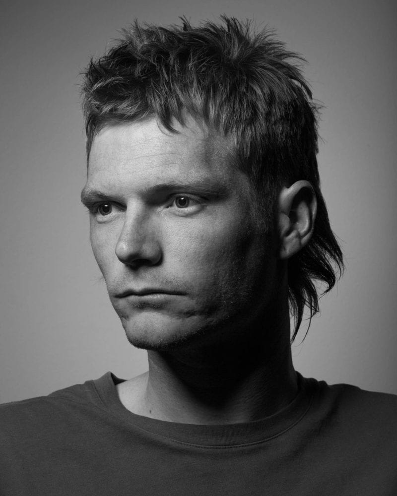 Man with a mullet in a black and white photo looks to the left of the camera without smiling