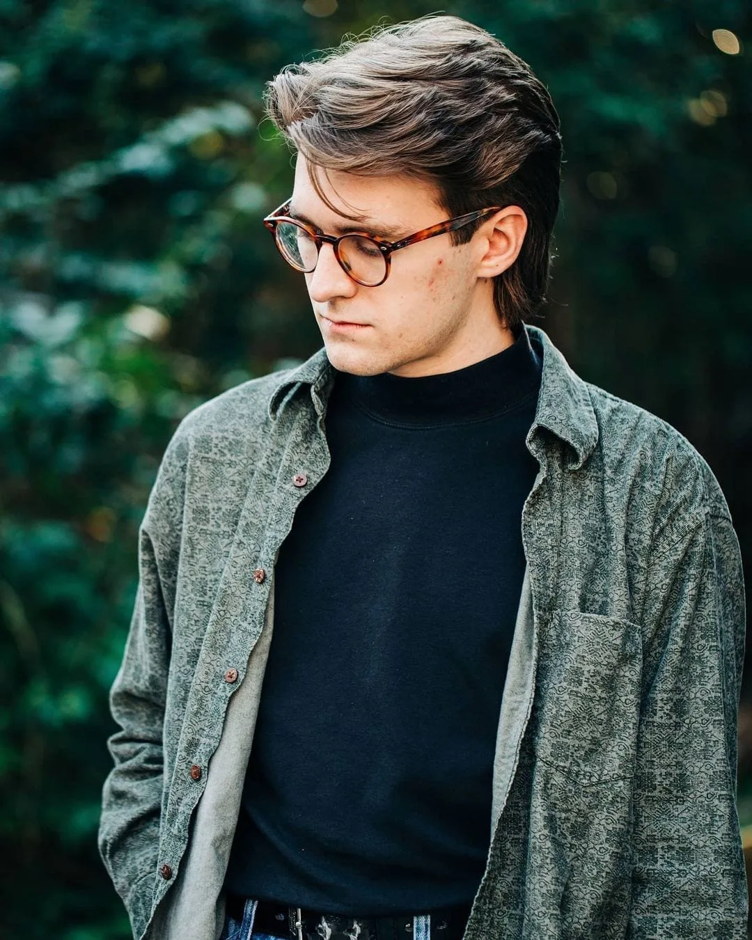 Modern hipster in a wool jacker and a turtleneck in glasses with a swept-back mullet looks down at the ground