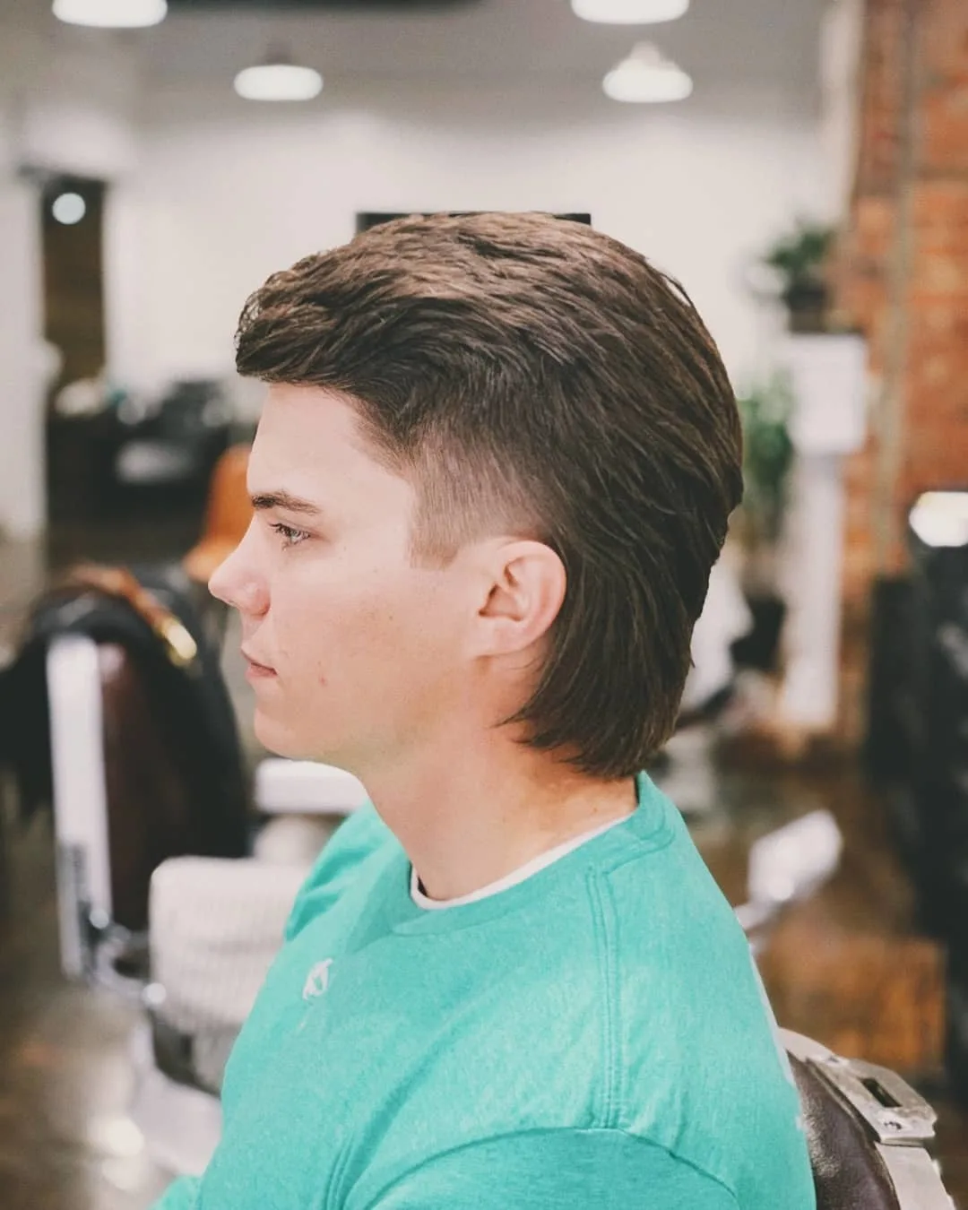 Guy in a green tshirt in a barber shop with a thick and faded mullet haircut