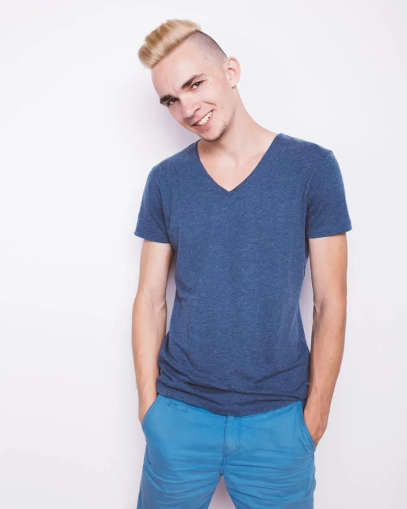 young guy with dyed hair stands near an old textured wall. street summer style in clothes: blue shirt and blue shorts. emotional portrait of a student. clean leather and short hair
