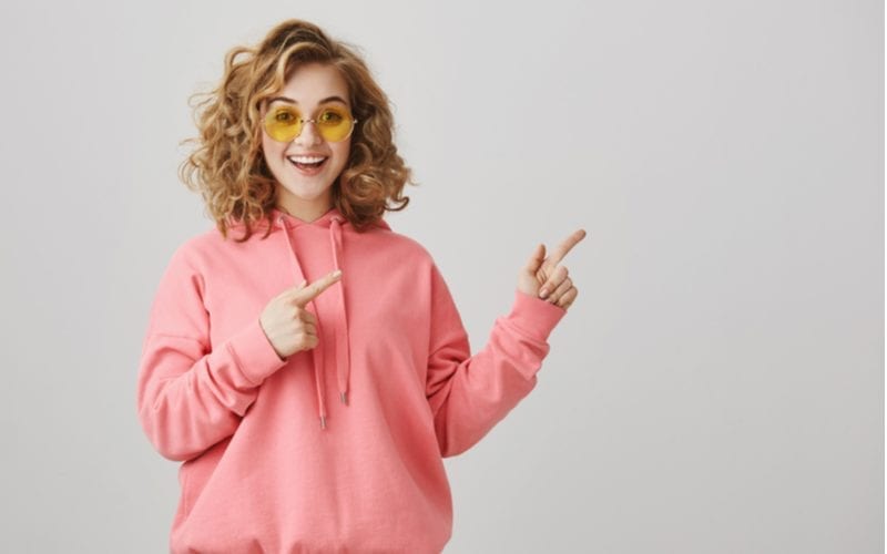 Shall we hit this bar and dance all night. Stylish attractive young woman with short curly hairstyle and trendy sunglasses pointing right with index fingers and smiling with interest over gray wall
