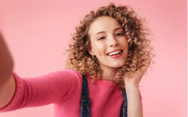 Portrait of happy young girl with curly hair taking a selfie isolated over pink background
