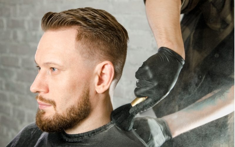 The barber cleans the neck of an adult man with a brush with talc after a haircut on a white brick wall background