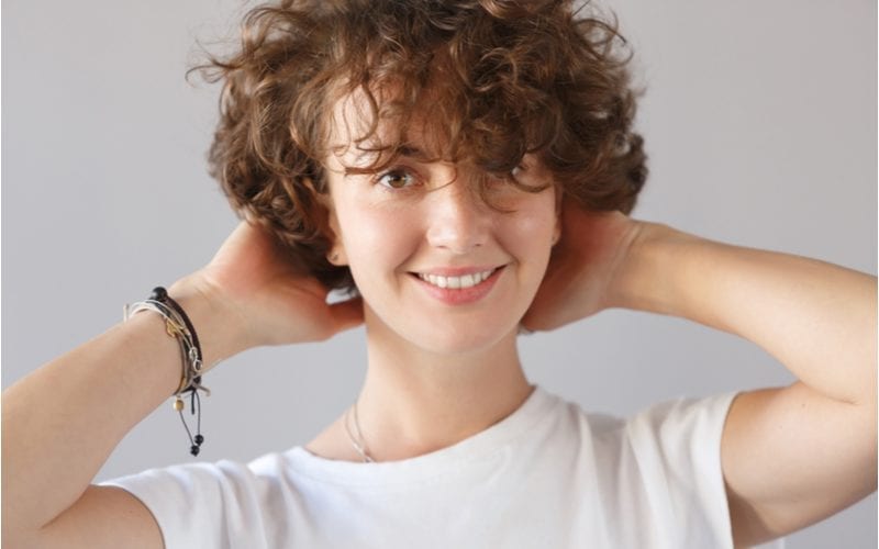 Cropped shot of beautiful happy funny female with curly hair and gentle smile, expresses positive emotions, poses in studioremoves his fancy bangs from his forehead