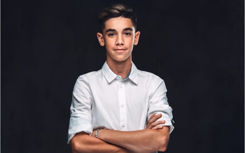 As an image for a piece on teen boy haircuts, young guy dressed in white shirt standing with crossed arms. Isolated on the dark background.