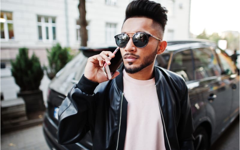 Stylish indian beard man at black leather jacket and sunglasses against business suv car. India model posed outdoor at streets of city and speaking on phone.