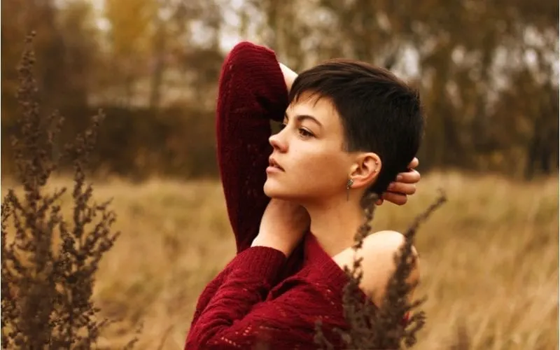 Girl with ultra short hairy pixie on nature on the field among dried flowers in autumn in a red knitted sweater