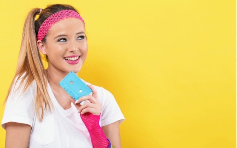 Against a yellow background is a woman holding a teal cassette tape with an unprofessional hairstyle of a side top ponytail
