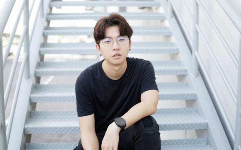 Asian man in glasses with a two block haircut sits on the steps of a building and looks at the camera