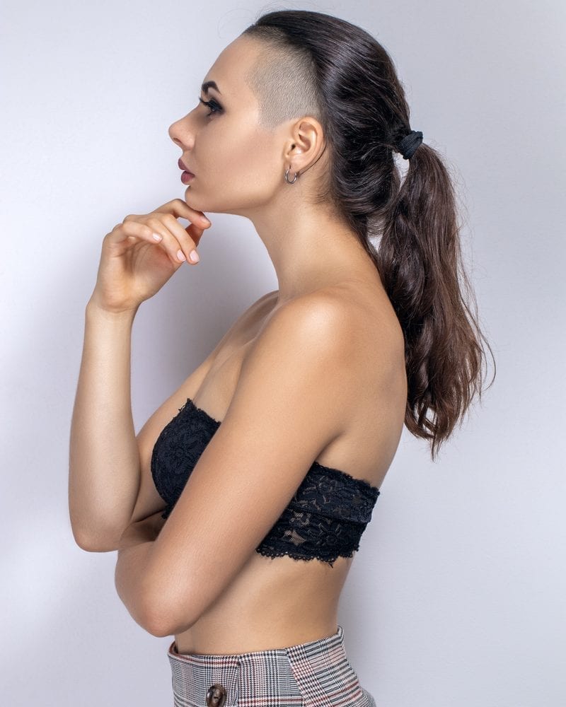 A woman with shaved head sides and a long back looks forward in a bra