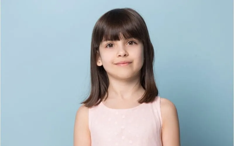 As a featured girl's haircut, a youngster stands in front of a blue background and grins at the camera