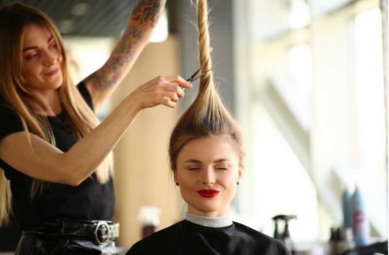 For a piece on short haircuts for women, a young woman closing her eyes while a stylist holds her long hair above her head and holds clippers up to the base of the pony