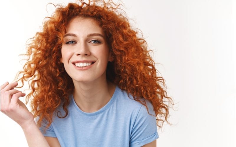 Alluring sensual young woman with natural curly red hair, rolling strand on finger silly, smiling toothy looking happy and coquettish, standing white background