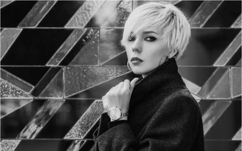 Close up monochrome, black and white fashion portrait of young beautiful woman with short blonde hair, smoky eyes makeup, wearing dark coat, elegant wrist watch. Copy, empty space for text