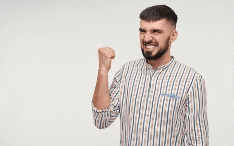 Pleased young pretty dark haired bearded male with short haircut showing happily his teeth and raising fist while looking aside, isolated over white background