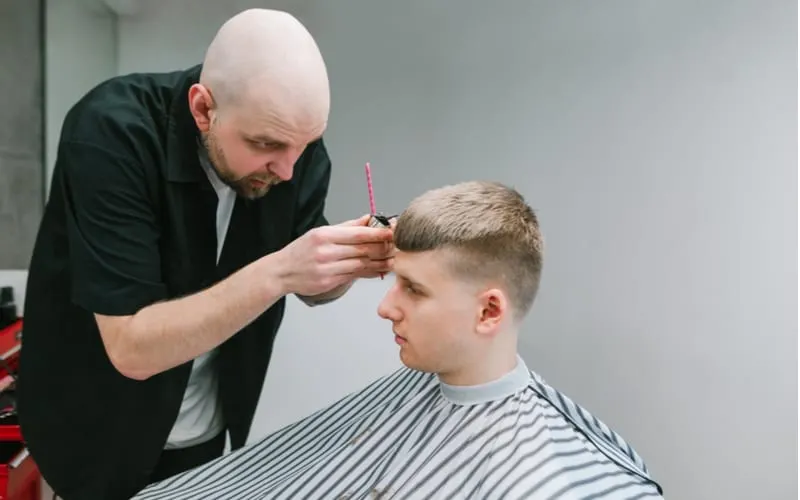 Barber with a trimmer in his hand creates a stylish hairstyle for the client of the barbershop, trims bangs. Serious male hairdresser uses a trimmer when cutting a client. French crop haircut.
