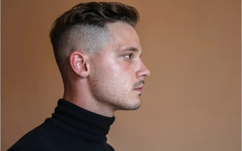 handsome young adult man with undercut hairstyle in profile view. Studio portrait of Man dressed in black turtleneck looking at camera against dark brown background. Shaved whiskey hairstyle