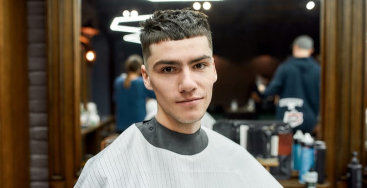 Portrait of a young caucasian guy with stylish haircut sitting in armchair in the modern barbershop or beauty salon and looking at camera