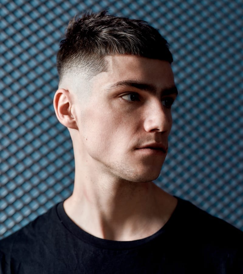 Portrait of a handsome young man in black T-shirt with stylish haircut looking away while posing against dark blue background. Beauty. Fashion. Mens haircut. Barbershop