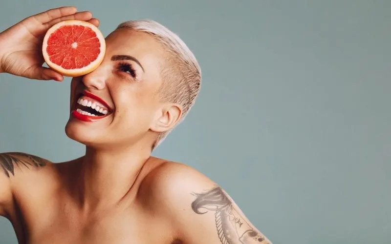 Close-up of a woman holding a grapefruit infront of her eye against grey background. Beautiful female model with short blond hair holding a grapefruit slice and smiling and rocking a Pixie Cut