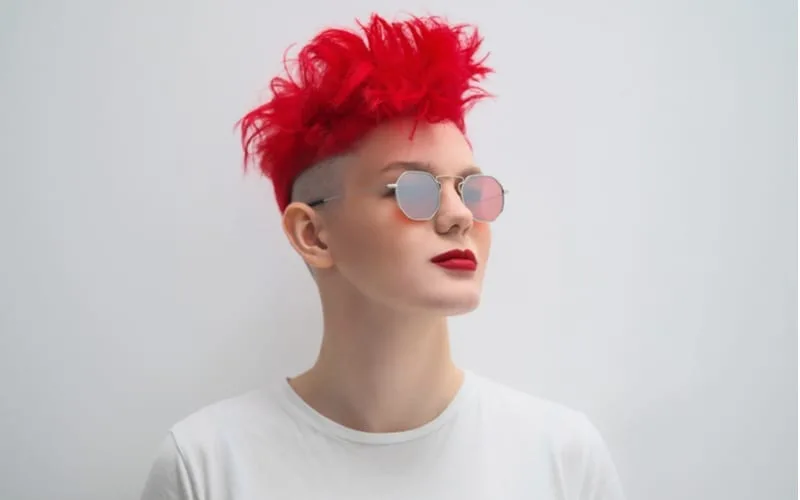 Portrait of a cool fashionable modern young girl. A short haircut with shaved temple. Dyed bright red hair. Red lipstick. Studio photo on a white background. Suspenders on checkered pants and