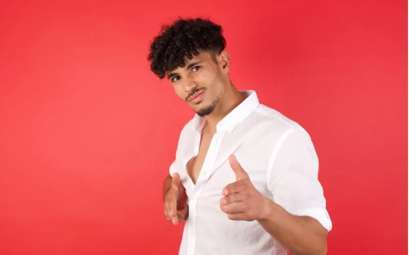 Joyful and charismatic good-looking emotive arab man with afro haircut winking and pointing with finger pistols at camera happily and cheeky posing over red wall