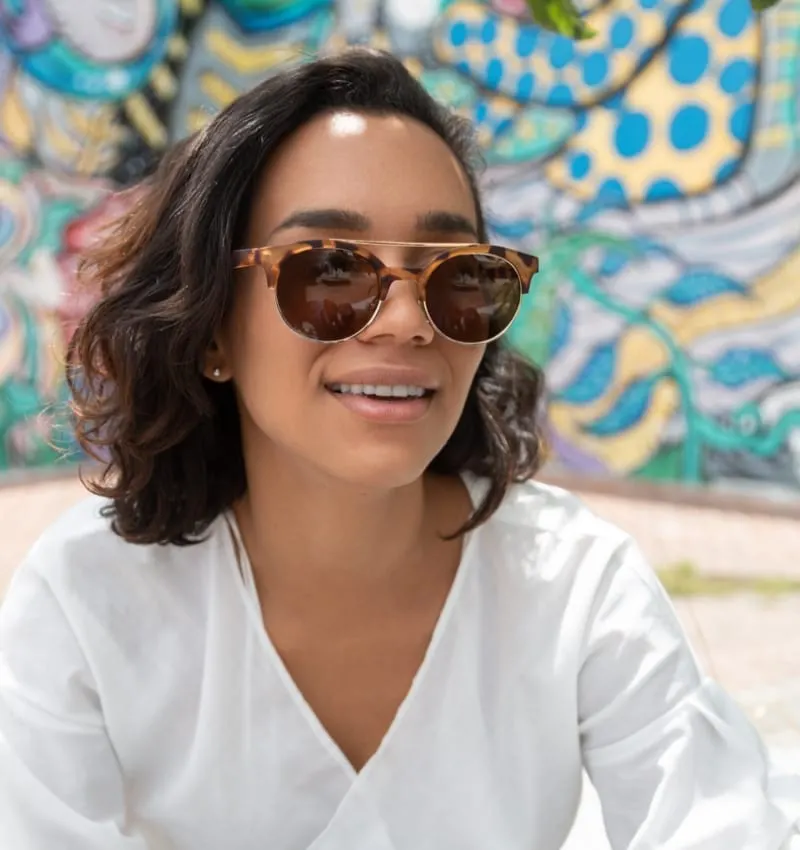 face of young brunette latina woman with short hair and curls, smiling and wearing sunglasses, she has a light white blouse, natural light with with colorful background outdoors