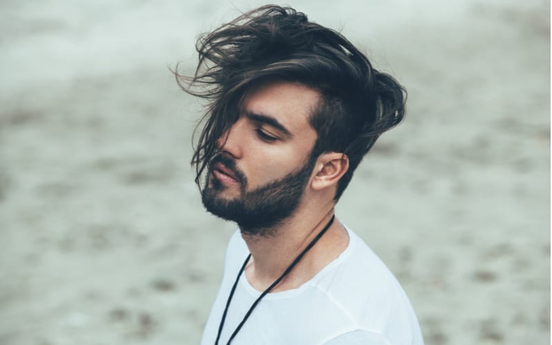 Man in a white tshirt with a long top short sides haircut with the long hair blown in his face on the beach