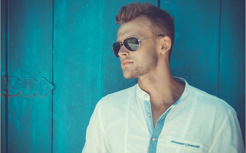 Portrait of a young with a high fade haircut and handsome man in a white shirt and trendy glasses posing over blue wooden background.