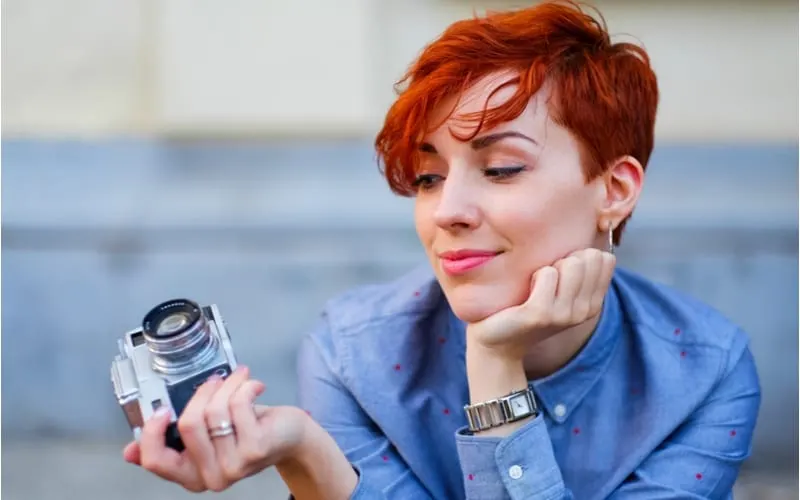 Beautiful smiling redhead woman with a short shag haircut in a denim shirt on red brick wall grunge, industrial background. Young stylish girl on the street with retro photo camera.