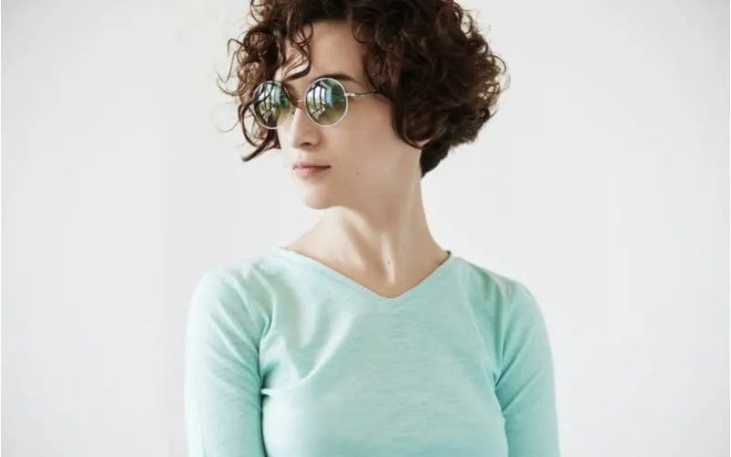 Portrait of young pretty woman with short curly shag hairstyle wearing round hipster sunglasses and mint top, looking away, posing against white studio wall background
