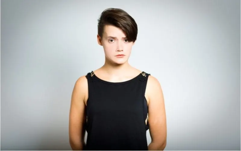 woman with a two block haircut holds her arms behind her back and frowns at the camera