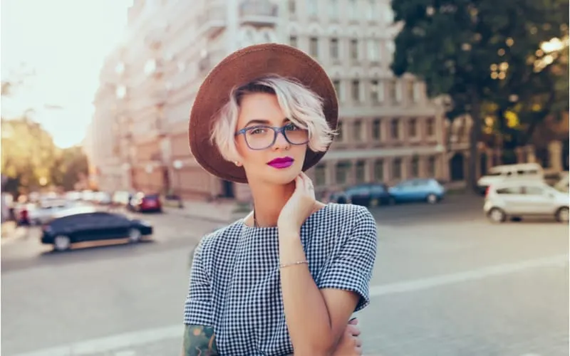 Portrait of pretty blonde girl with short hair posing to the camera on the street in city. She wears gray checkered dress, glasses, hat and has purple lips.