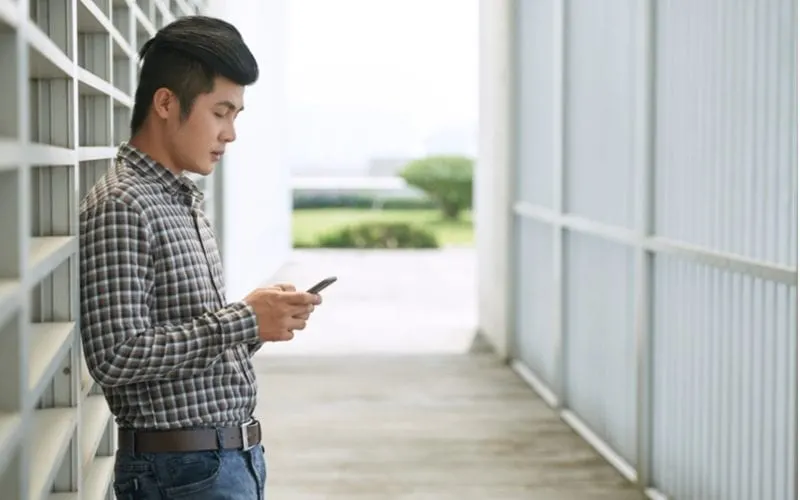 Side profile of a young asian man standing in a warehouse with his shirt tucked in and looking at a phone