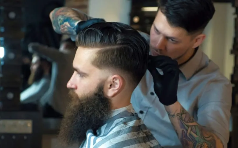 A guy with a short sides long top haircut is sitting by a handsome bearded man is looking forward while having his hair cut by hairdresser at the barbershop