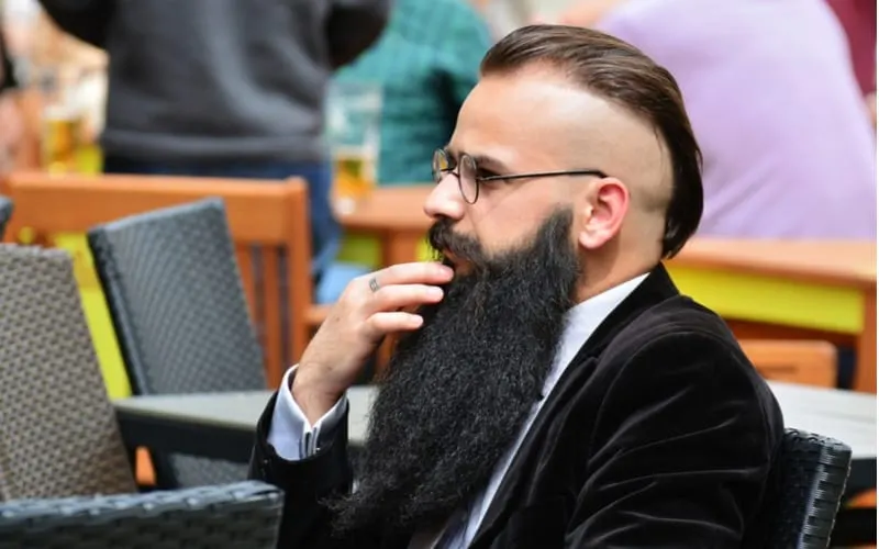 Elegant middle aged business man sits at a street cafe table with a modern slicked back disconnected undercut hairstyle with shaved sides and a long black beard.