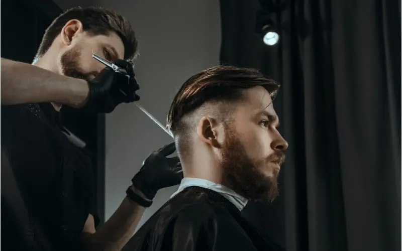 BARBERSHOP THEME. BEARDED BARBER IN BLACK RUBBER GLOVES IS TRIMMING THE HAIRCUT OF HIS YOUNG SERIOUS CLIENT. HE IS USING A HAIR CLIPPER