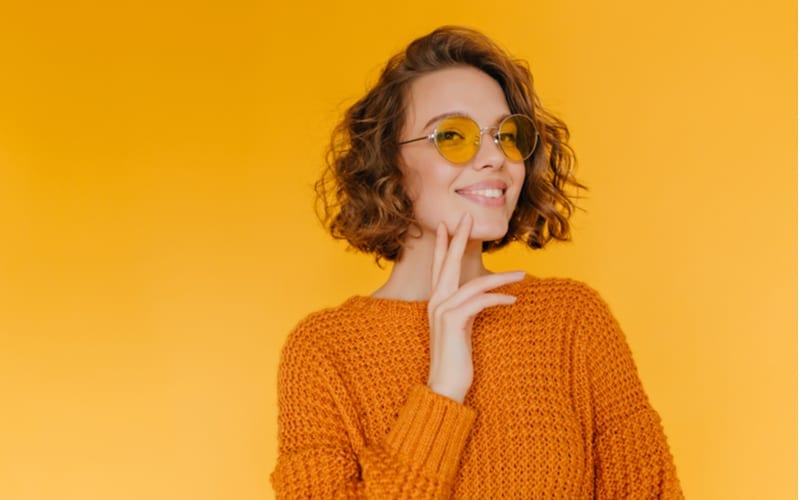 For a piece on short shag haircuts, Cheerful short-haired girl in trendy glasses posing with pleasure in new knitted attire. Indoor portrait of pleased curly young woman standing in front of yellow wall.