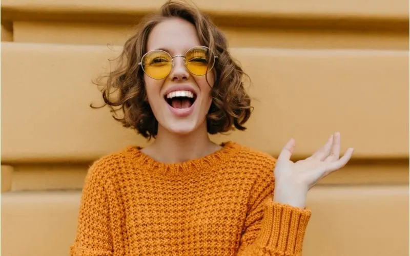 Enthusiastic smiling girl with shiny curly haircut posing in front of old wall. Close-up outdoor portrait of enchanting lady in sweater and trendy glasses.