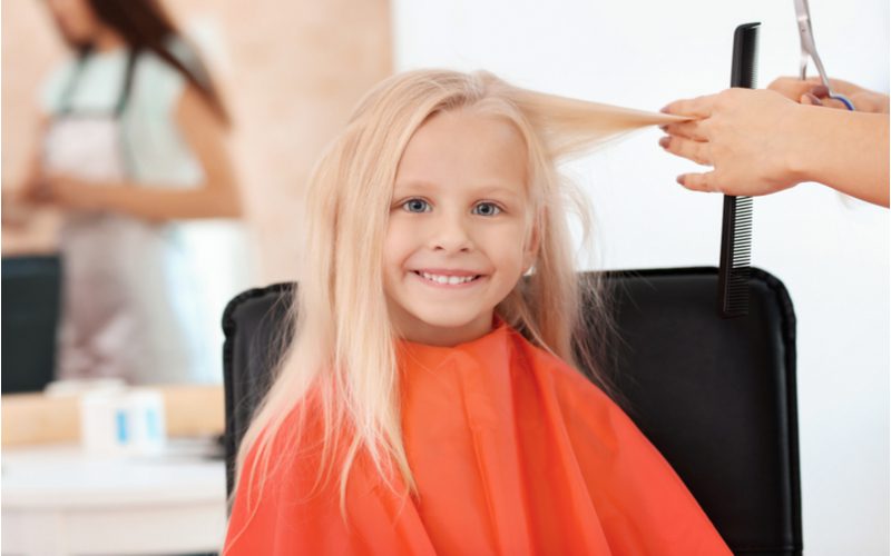 Image for a piece on little girl haircuts featuring a young girl in a red cape getting her blonde hair cut while she's smiling in a salon chair