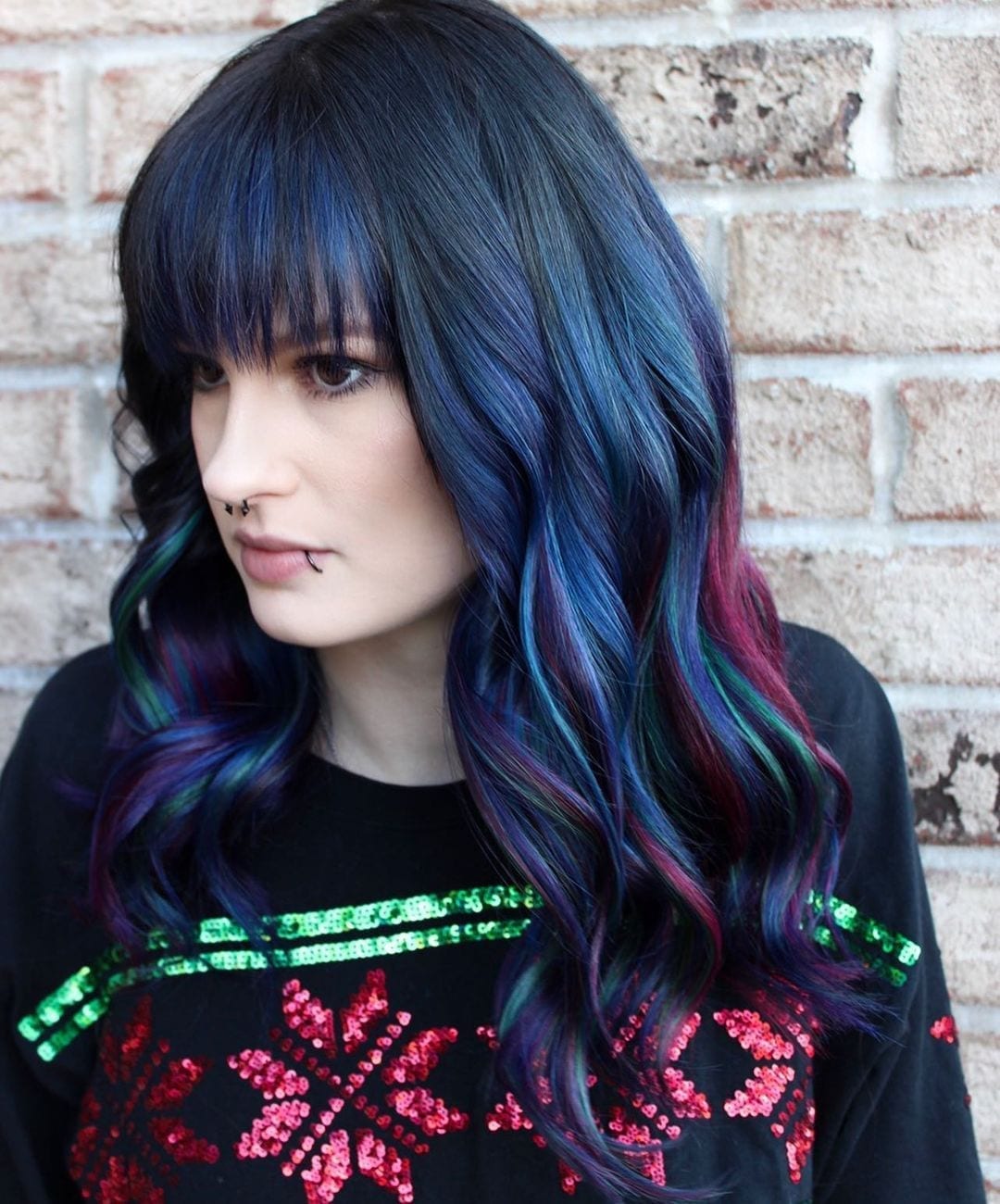 Pale woman with a lip ring and dark blue hair does not smile and wears and black and multi-colored sweater