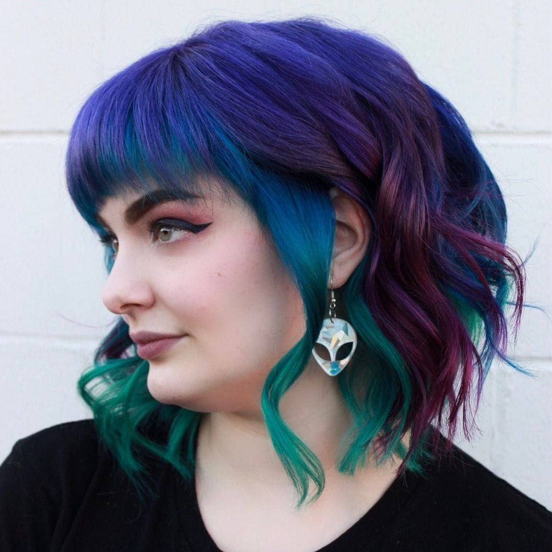 Person wearing alien earrings and with blue hair looks right and does not smile
