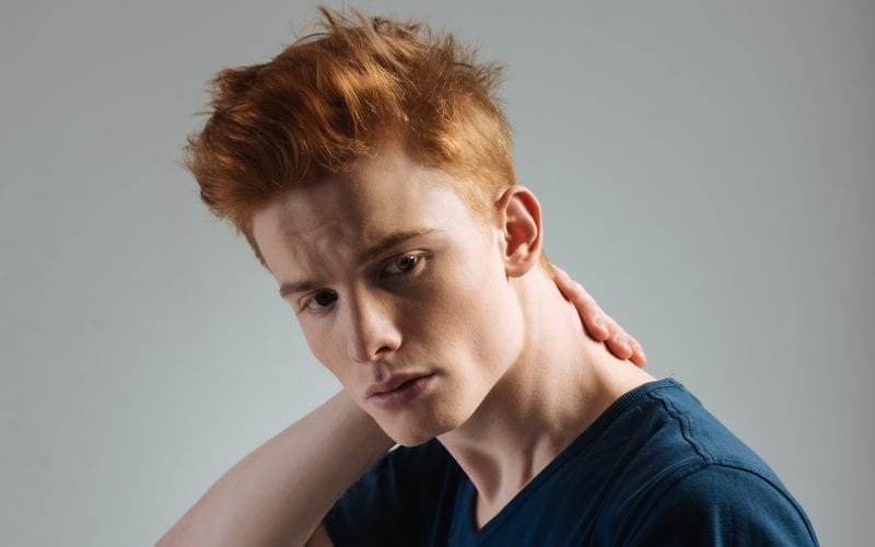 Man wearing a red headed hairstyle holds the back of his neck and looks below the camera while standing in a studio