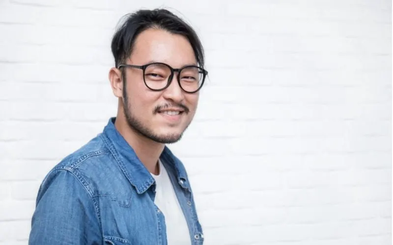 Hipster asian man with a parted bowl cut stands against a block background and wears an open jean jacket with white tshirt