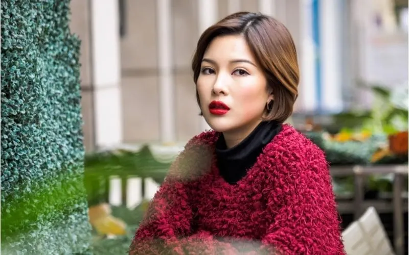 Well dressed woman with a red sweater and red lips sits in front of vegetation