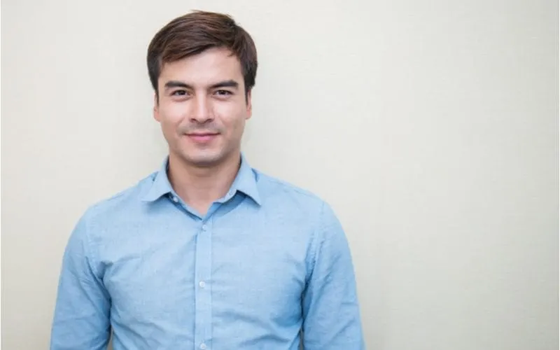 Young man with a blue button up shirt grins and looks intensely at the camera for a piece on best haircuts for men with widows peak